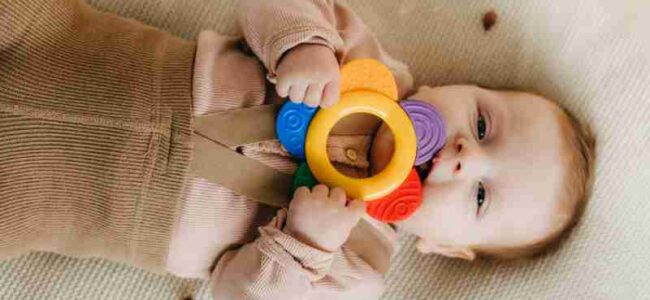 Cute baby chewing on a colorful teething toy while lying on a soft surface., tags: eltern wegen erstickungsgefahr nicht - pexel